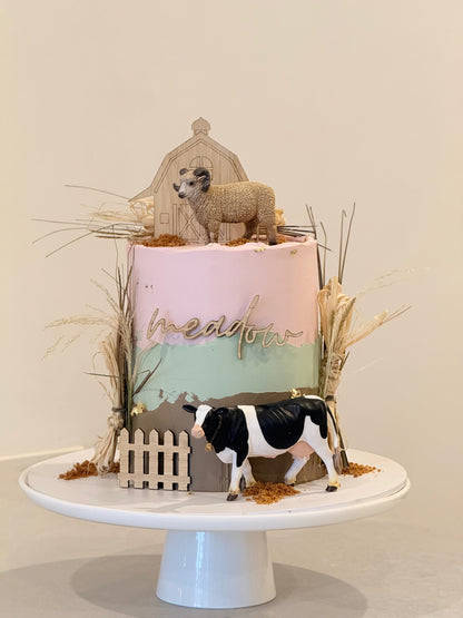 Decorative cake with sheep and cow figurines on a white stand against a beige background