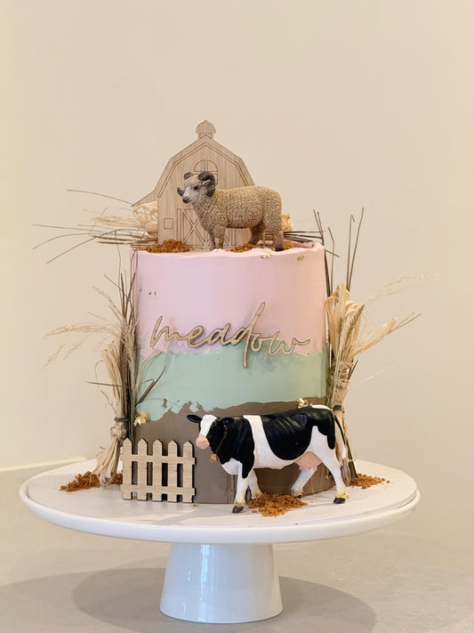 Decorative cake with sheep and cow figurines on a white stand against a beige background