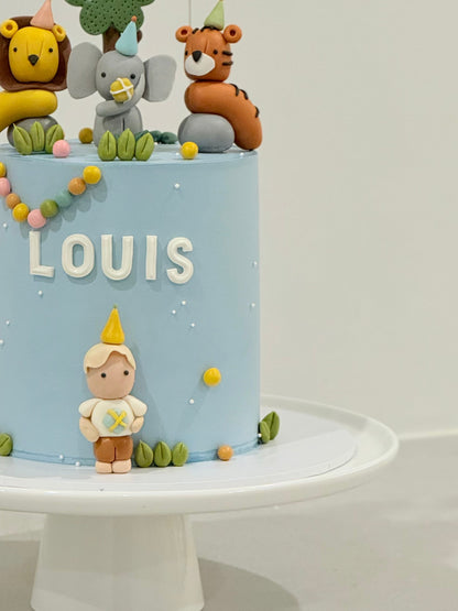 Children's birthday cake with animal figurines and 'LOUIS' on a light blue background