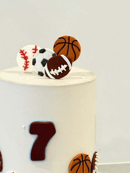 Sports-themed cake toppers including a basketball, football, baseball, and soccer ball on a white cake.