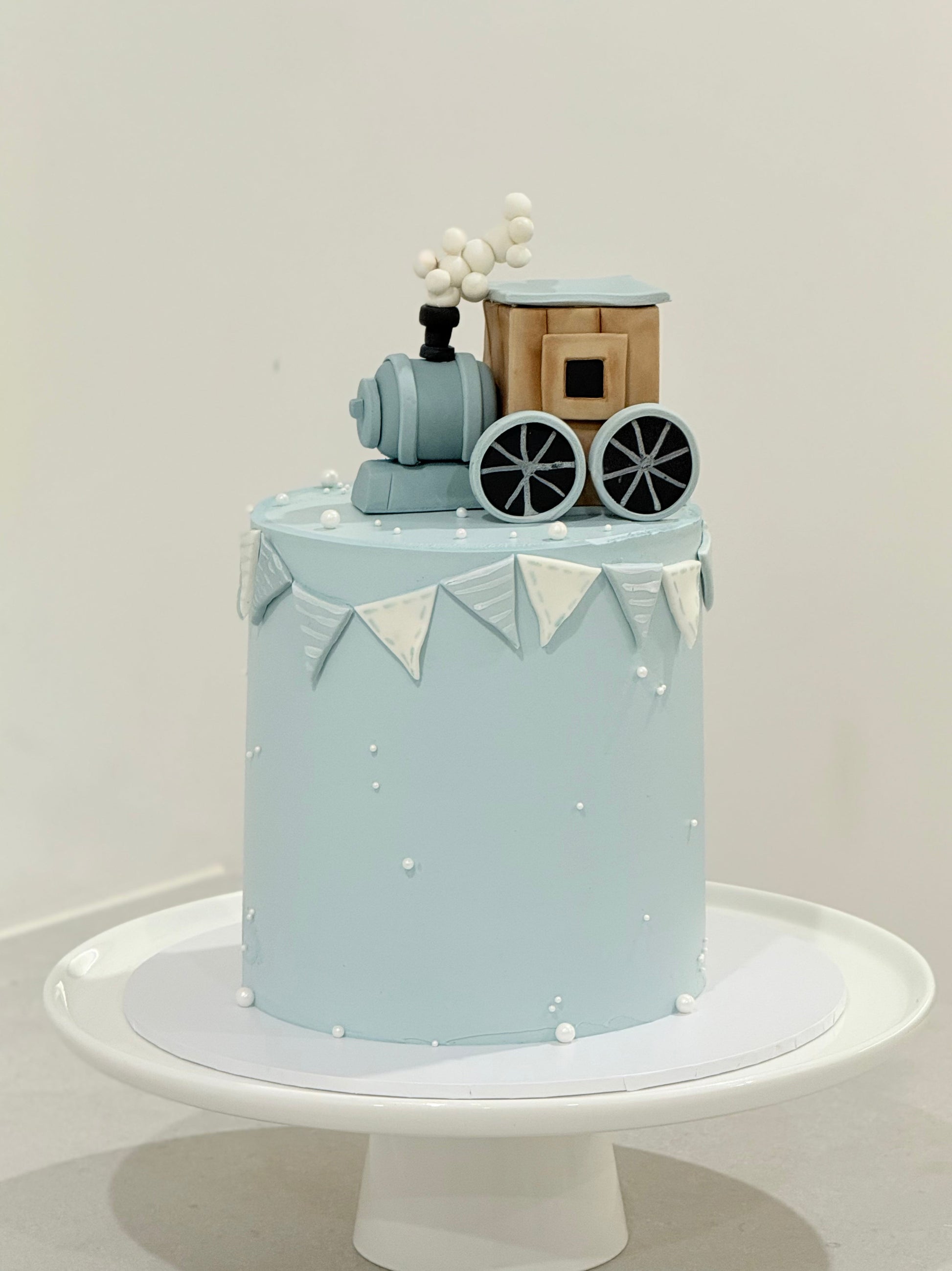 Light blue cake with train decoration on a white stand against a light gray background