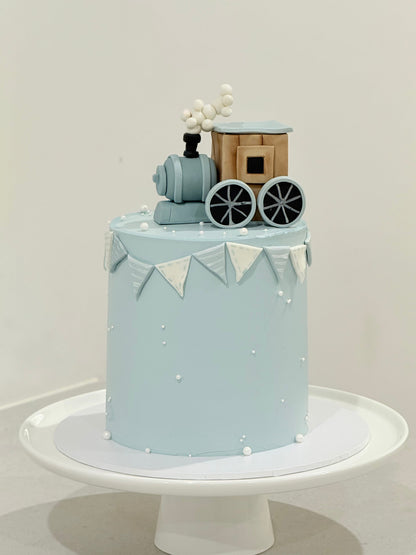 Light blue cake with train decoration on a white stand against a light gray background